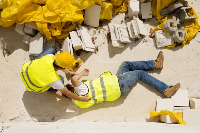 A construction man on the floor being put into a recovery position by a colleague after a working at height accident using bad fall restraint systems.
